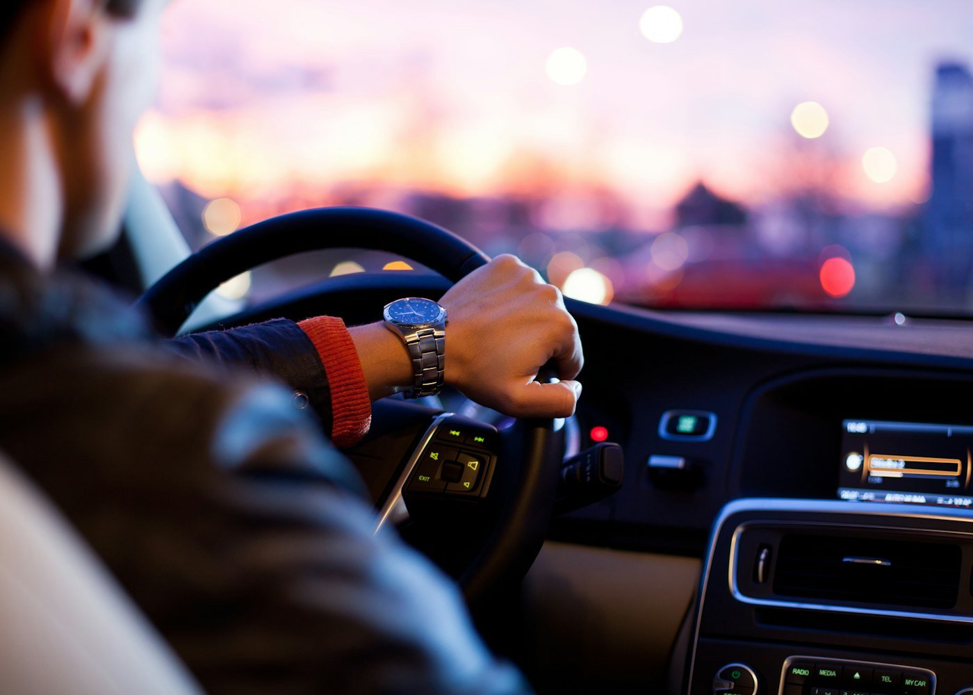 View from the driver's seat of a car at dusk, hands near the steering wheel and lit dashboard
